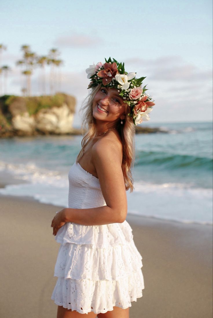 a beautiful young woman standing on top of a beach next to the ocean wearing a flower crown