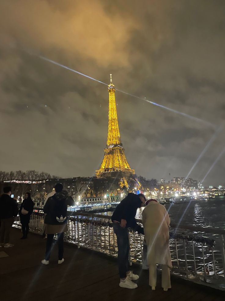 people looking at the eiffel tower lit up in yellow and white from across the river