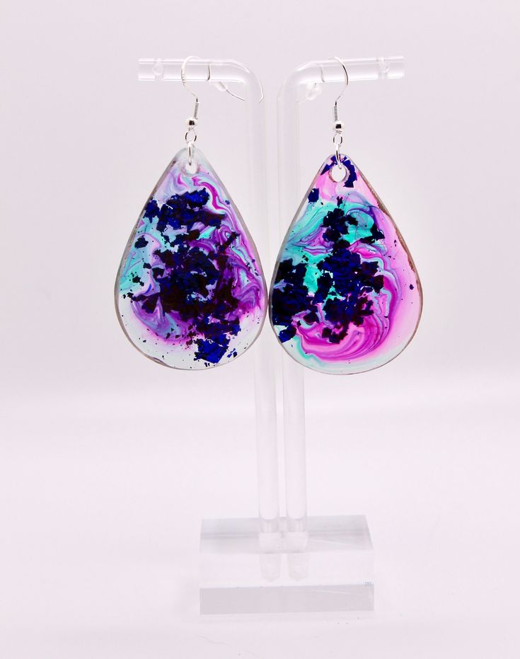 a pair of earrings with purple and blue paint on them, hanging from a clear acrylic stand