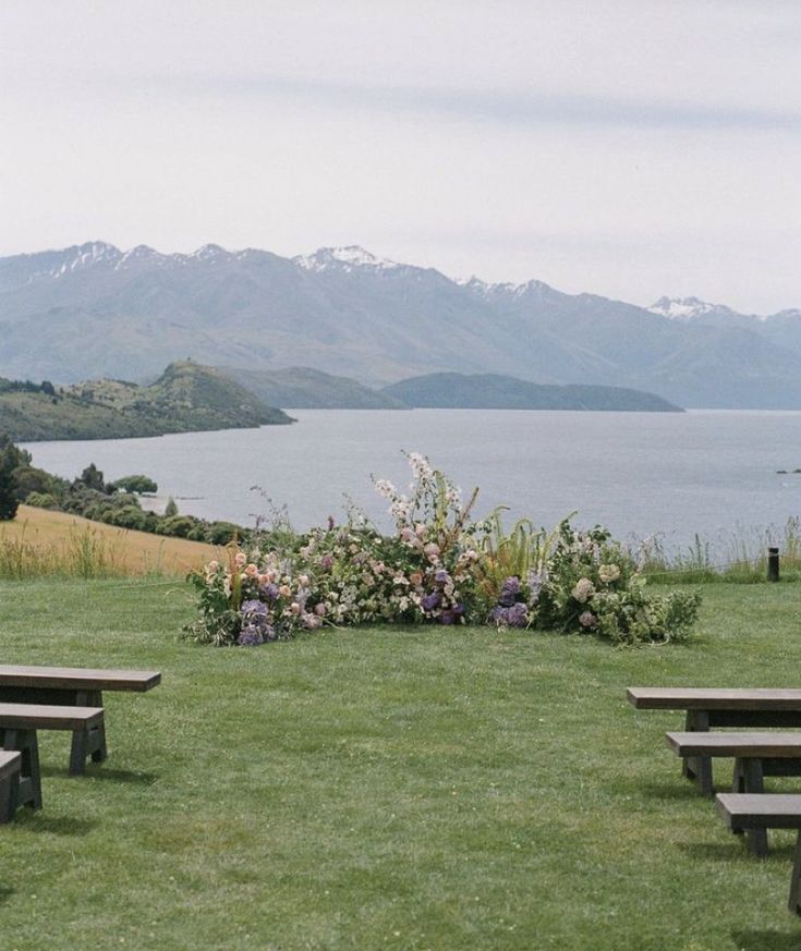 there are two benches in the grass by the water and mountains behind them, with flowers growing on the lawn