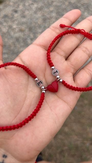 a hand holding two red string bracelets with silver beads and heart charms on them