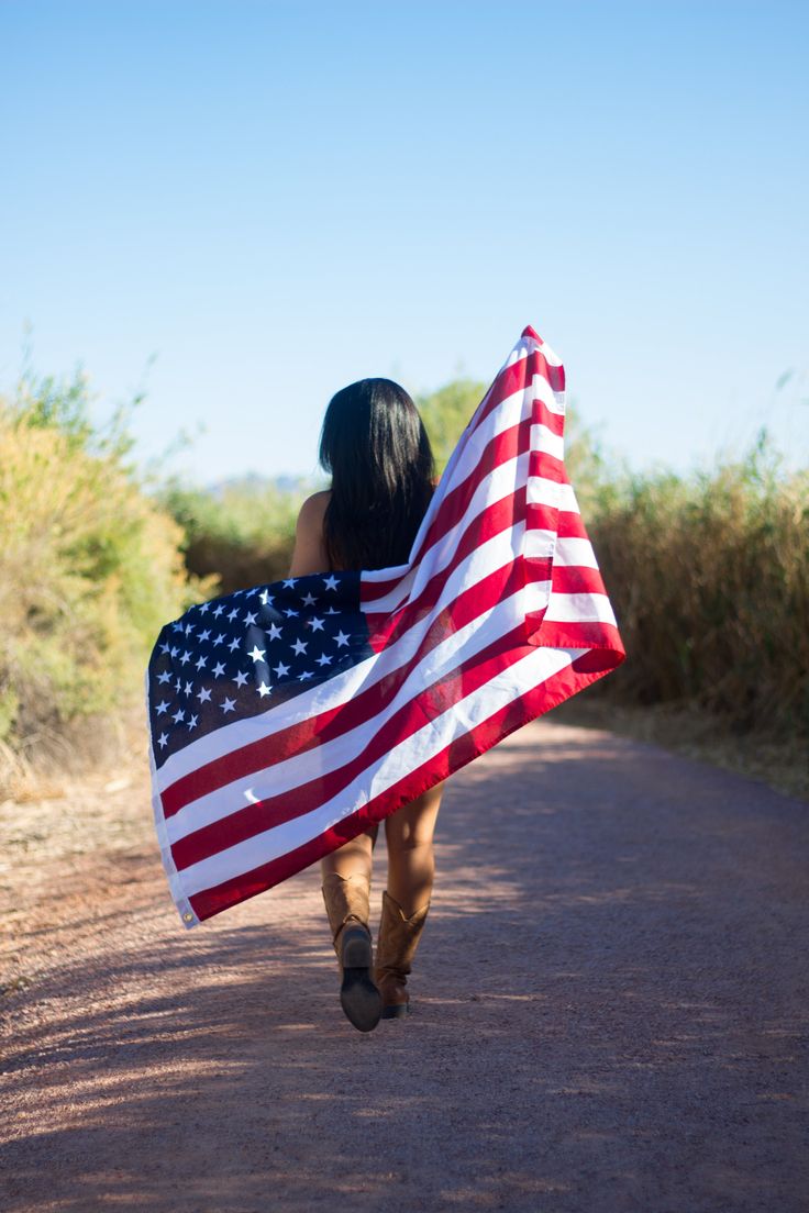 a woman walking down a dirt road carrying an american flag on her back and covering her face