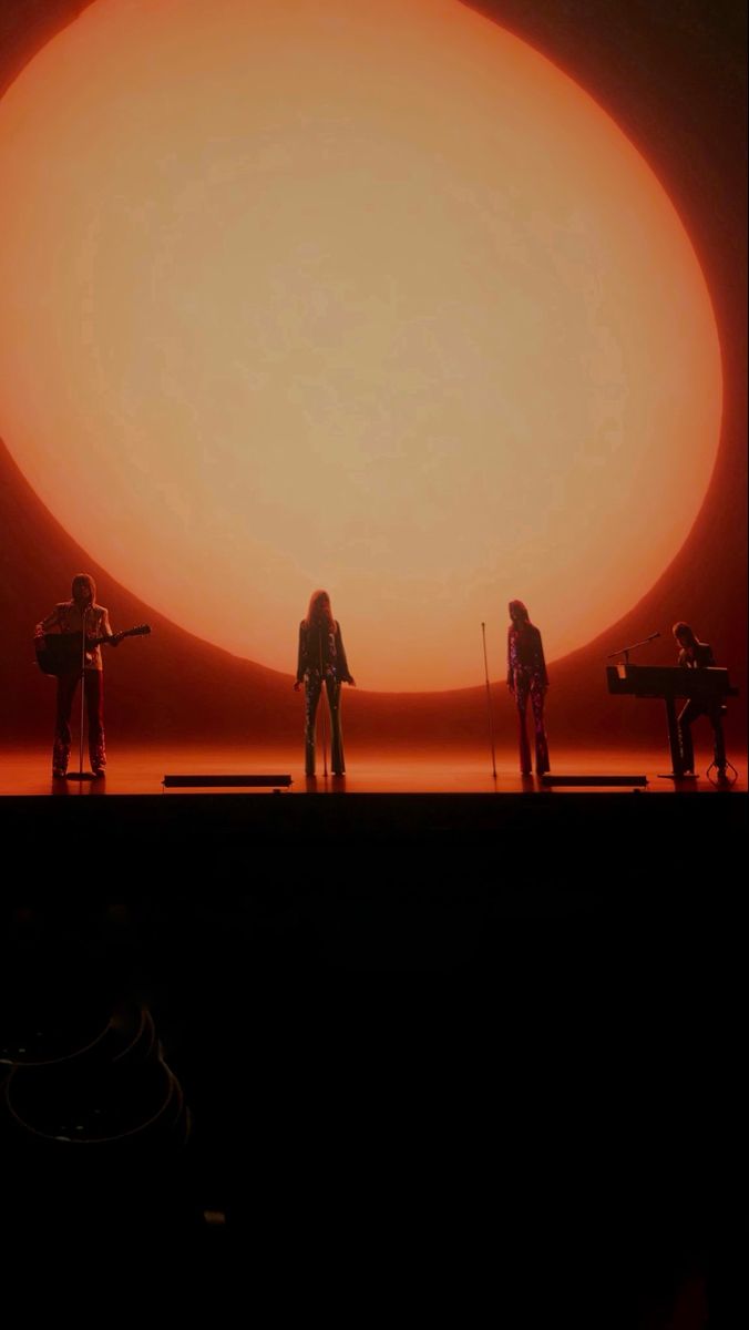 three people standing on stage in front of an orange sun with one person holding a microphone