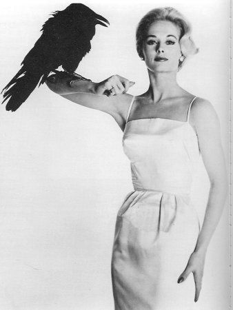 a woman in a white dress holding a black bird on her arm and looking at the camera