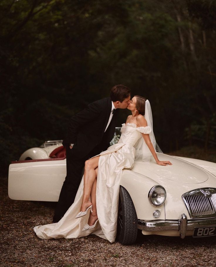 a bride and groom kissing in front of an old fashioned white car on the road