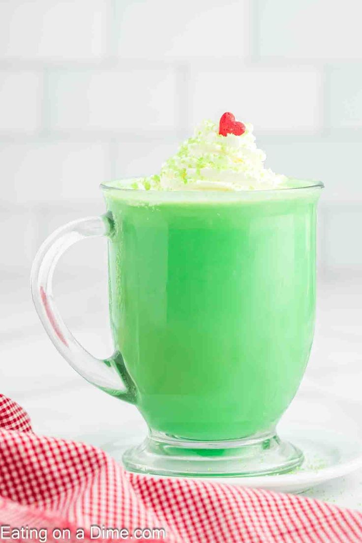 a green drink with whipped cream and a cherry on top in a glass mug sitting on a red checkered napkin