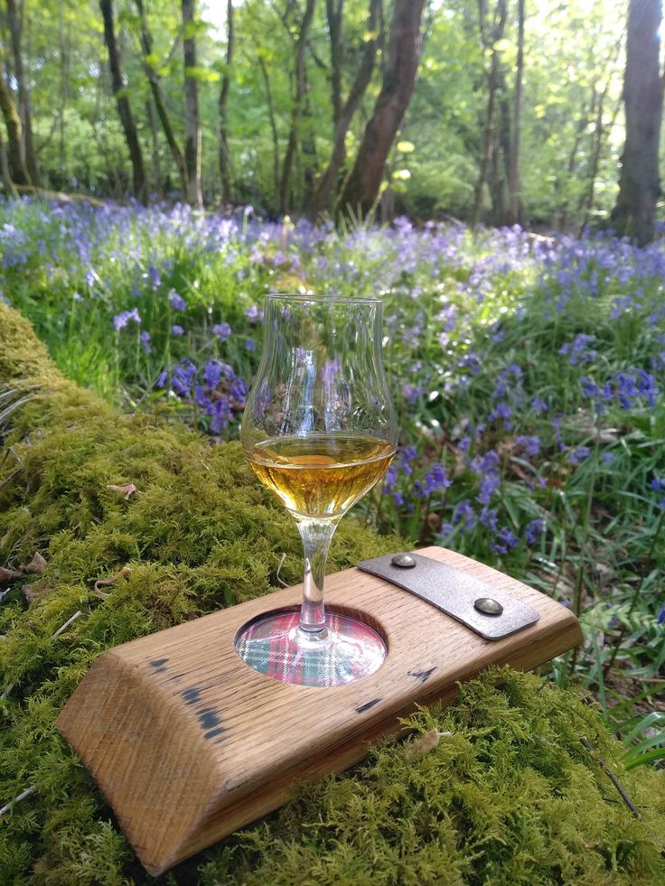 a glass of wine sitting on top of a wooden tray in the woods next to bluebells