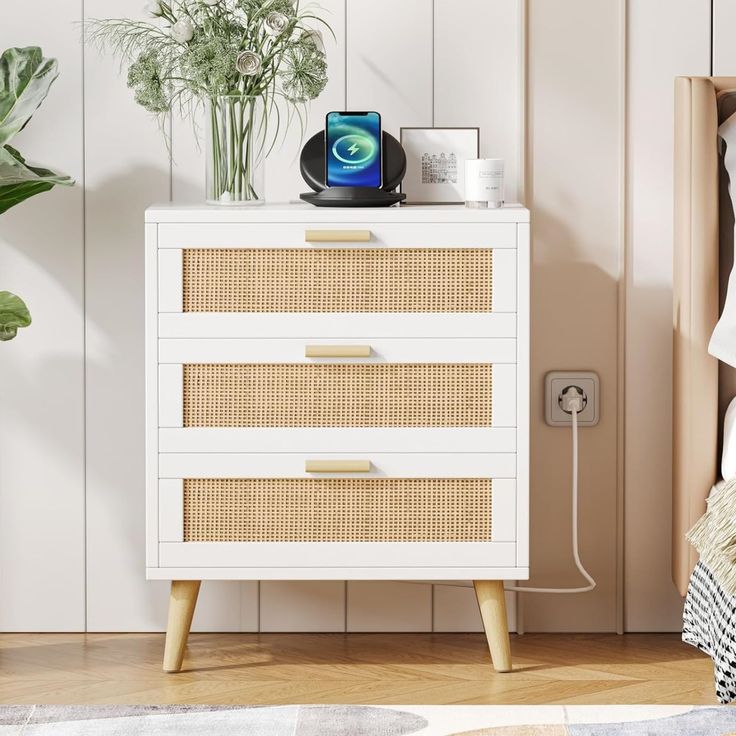 a white dresser with wicker drawers next to a plant and phone charger on top