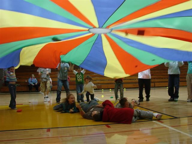 a group of people standing and sitting around a large colorful umbrella on top of a basketball court