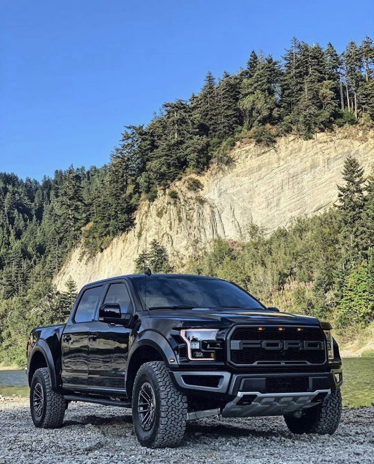 a black truck parked on top of a gravel field next to a forest covered hillside