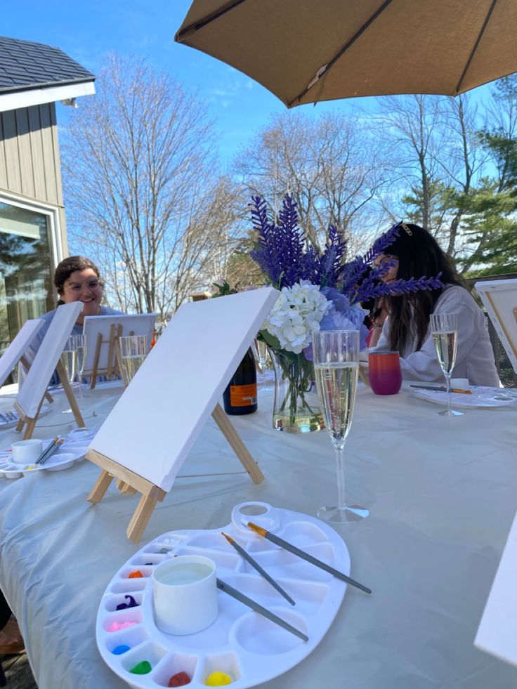 two women sitting at an outdoor table with art supplies and paintbrushes on it