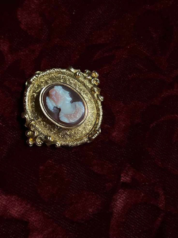 An adornment worthy of a museum! This stunning 19th century Sardonyx Cameo is a testament to Napoleon era French jewelry design. Crafted circa 1850-1860 in 18 carat gold, this brooch features a vibrant cameo of a aristocratic woman, set again a delicate peachy-hued sardonyx mount. The 18-carat gold mounting is also exceptional, with a gilded design brimming with fine detailing, and scrolling foliate motifs. The silhouette it creates makes it feel as if it should be hung in the halls of a palace! Formal Gold Cameo Brooch, Formal Gold Cameo Brooches, Elegant Gold Cameo Brooch, Ornate Gold Cabochon Brooches, Heirloom Cabochon Brooches For Formal Occasions, Heirloom Brooches With Cabochon For Formal Occasions, Victorian Cameo Brooches In Yellow Gold, Victorian Yellow Gold Cameo Brooches, Victorian Cameo Brooch In Yellow Gold