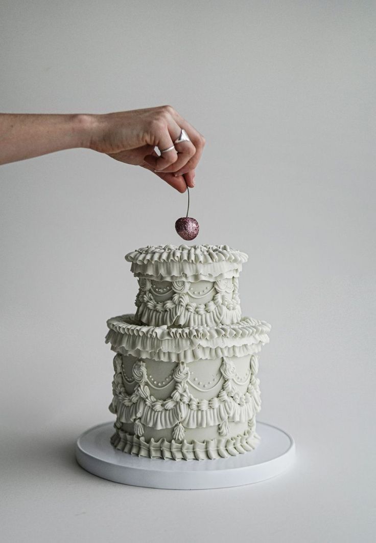 a hand is reaching for a cherry on top of a tiered cake with white icing