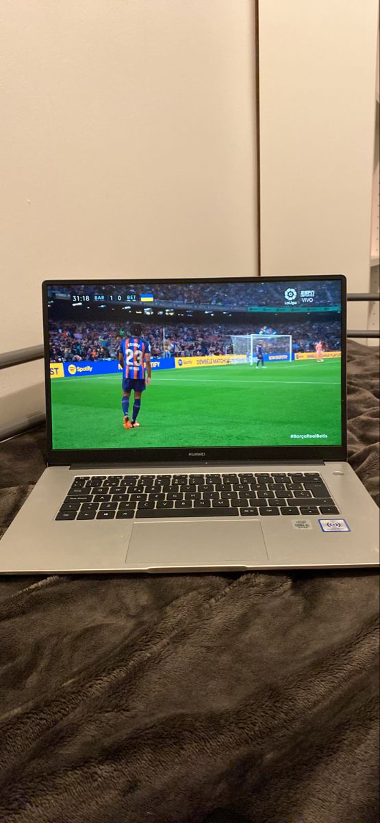 an open laptop computer sitting on top of a bed next to a wall with a soccer game on it