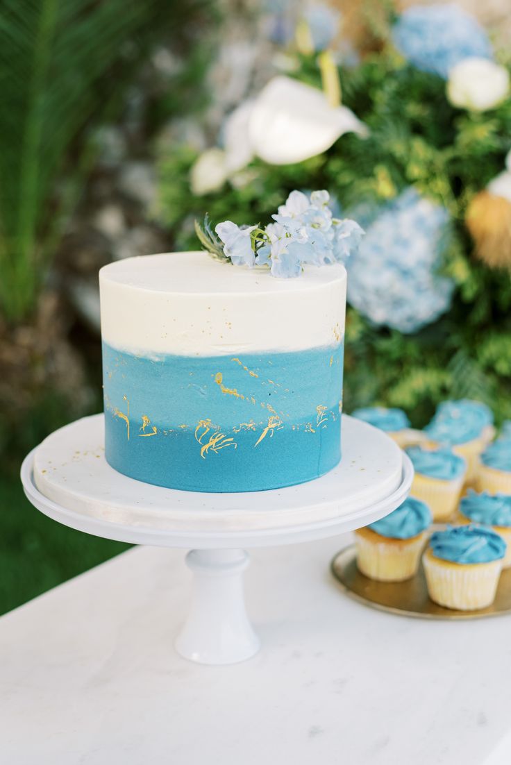 a blue and white cake sitting on top of a table next to cupcakes