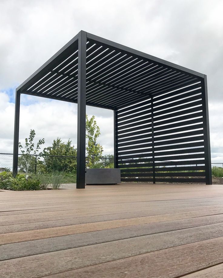 an empty wooden deck with a black pergolated structure in the middle and trees in the background