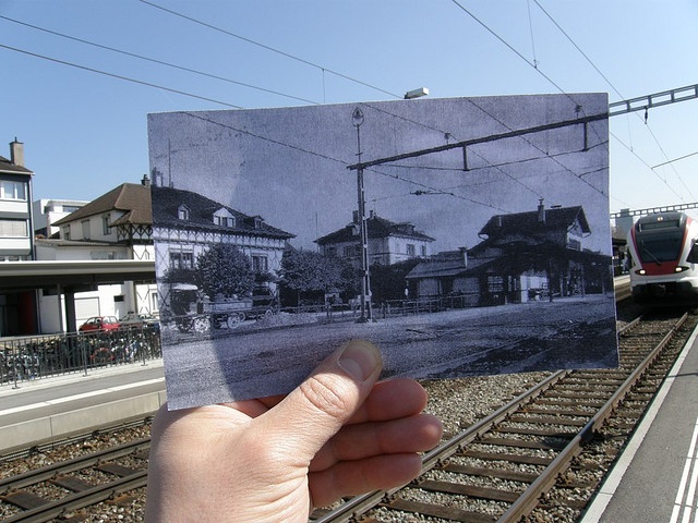 a hand holding up an old photo in front of a train track with houses on the other side