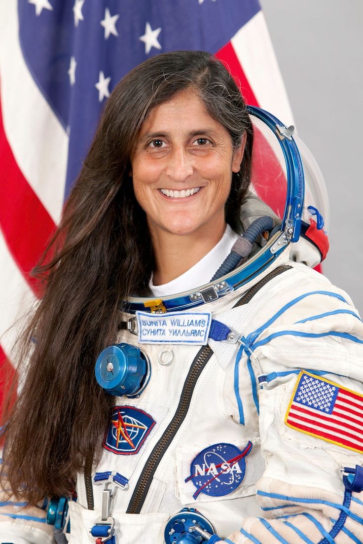 a woman in an astronaut's suit standing next to the american flag and smiling