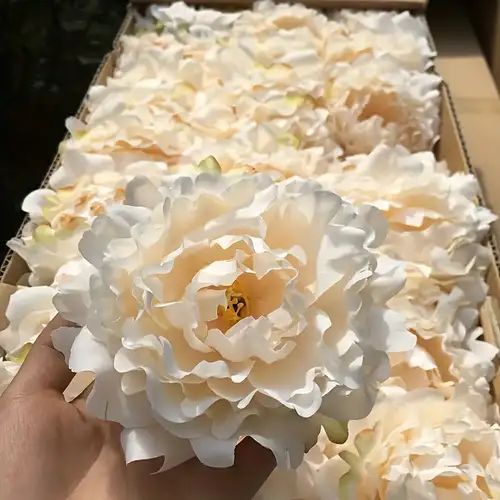 a person holding a large white flower in front of a box filled with fake flowers