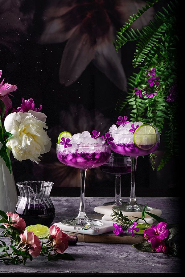 two glasses filled with purple and white liquid next to flowers on a wooden table in front of a dark background