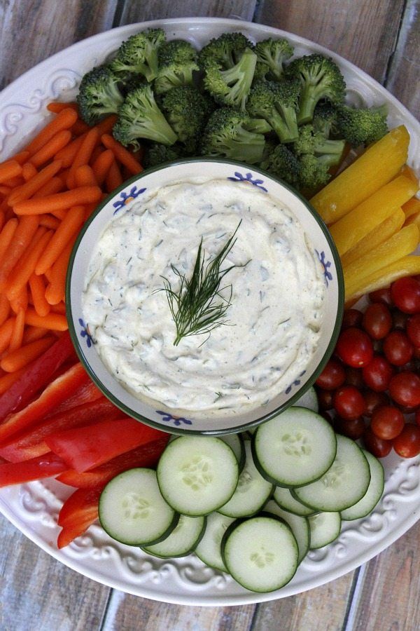 a white plate topped with veggies and dip surrounded by carrots, cucumbers, broccoli