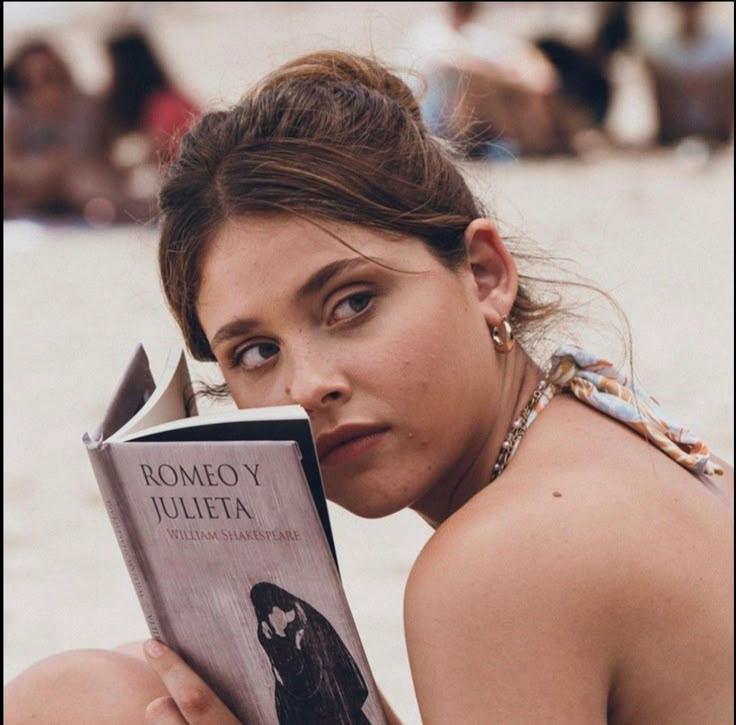 a woman sitting on the beach reading a book with her eyes closed and looking at the camera