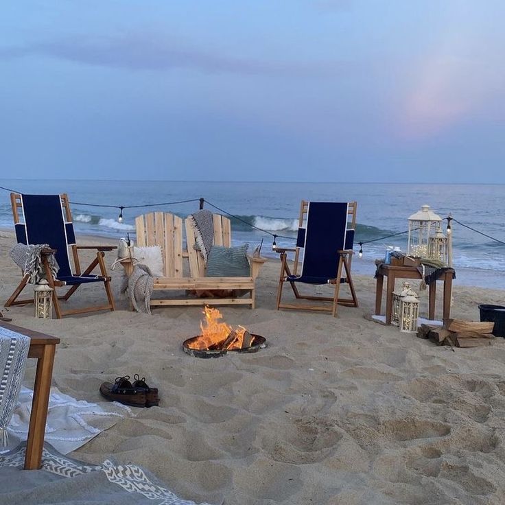 a fire pit sitting on top of a sandy beach next to the ocean with chairs