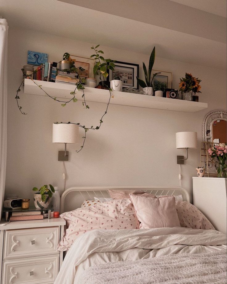 a white bed sitting under a window next to two lamps and a shelf filled with plants