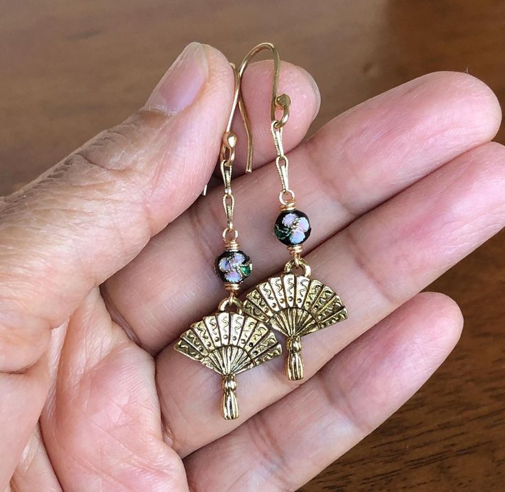 These charming gold fan earrings feature small round cloisonne beads with elegant gold-colored fan-shaped charms. Completed with gold-colored earring wires, these earrings are lightweight and comfortable. Whether you're dressing up for a special occasion, adding a unique touch to your everyday look, or searching for a thoughtful gift for someone special, they are sure to make a lasting impression. Nickel Free Enamel Dangle Earrings, Gift Brass Beaded Earrings With Round Beads, Gold Beaded Earrings With French Hook As Gift, Gold Jewelry With French Hook And Round Beads, Unique Brass Bead Shaped Earrings, Unique Brass Beaded Earrings, Unique Brass Earrings With Round Beads, Brass Beaded Round Earrings For Gift, Round Brass Beaded Earrings For Gift