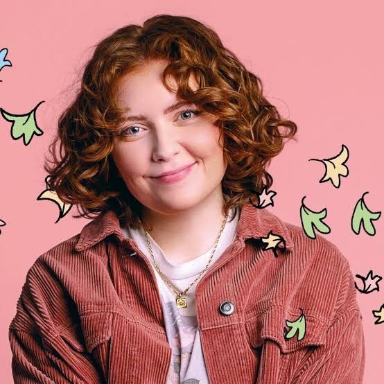 a woman with curly hair wearing a red jacket and smiling at the camera while standing in front of a pink background