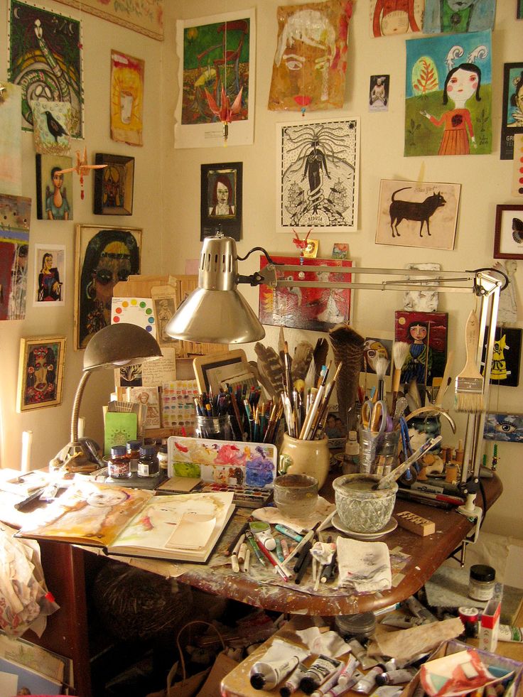 a cluttered desk with lots of art work on the wall and pictures all around it