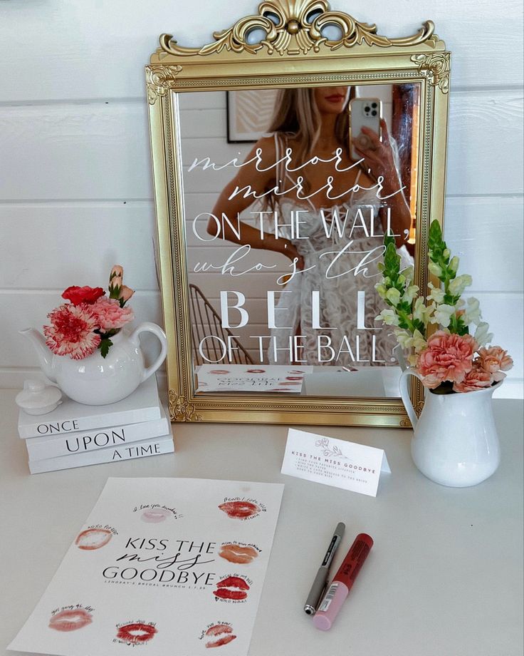 a table topped with a mirror and vase filled with pink flowers next to a sign