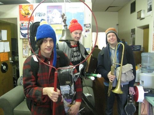 three men are standing in an office with instruments and hats on their heads, one man is holding a cell phone to his ear