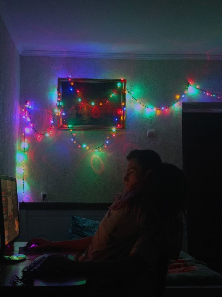 a man sitting in front of a computer with christmas lights on the wall behind him