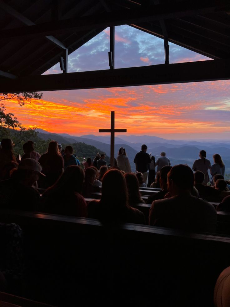 a group of people sitting in front of a cross on top of a hill at sunset