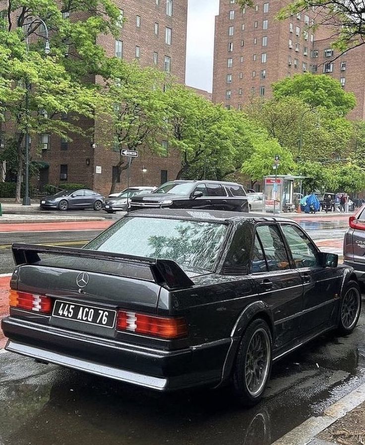 a black car parked on the side of a wet street next to tall buildings and trees