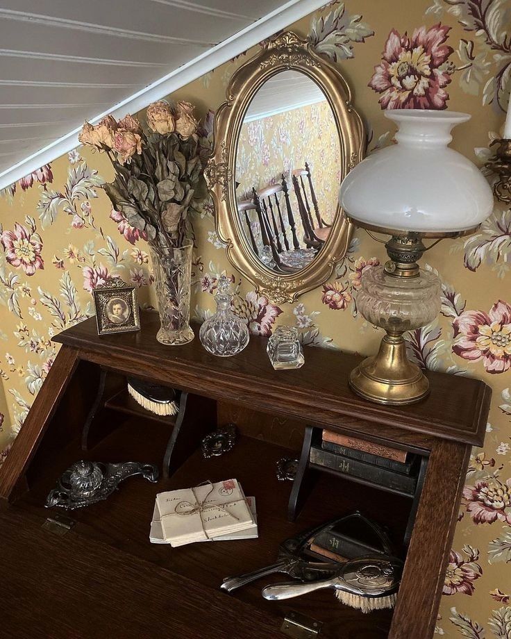 a desk with a mirror, vases and other items on it in front of a floral wallpaper