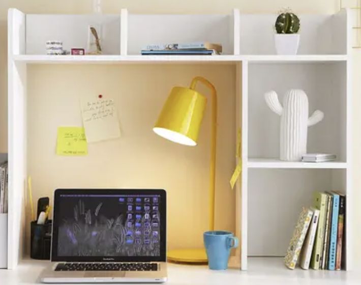 a laptop computer sitting on top of a white desk next to a lamp and bookshelf