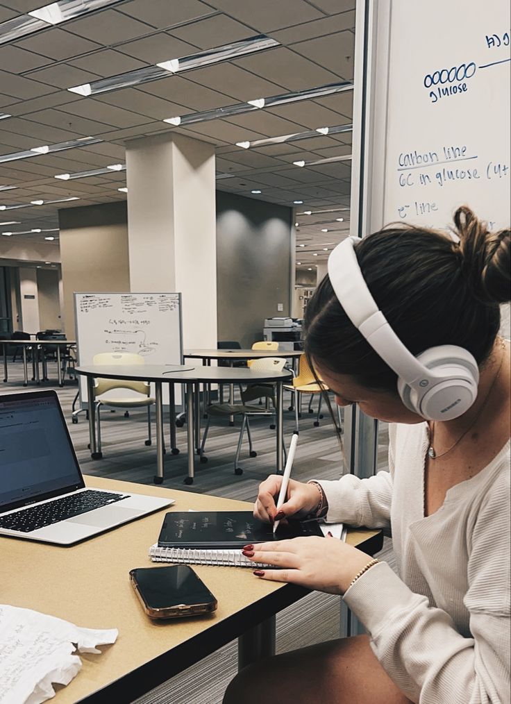 a woman sitting at a desk with headphones on, writing in her notebook and using a laptop