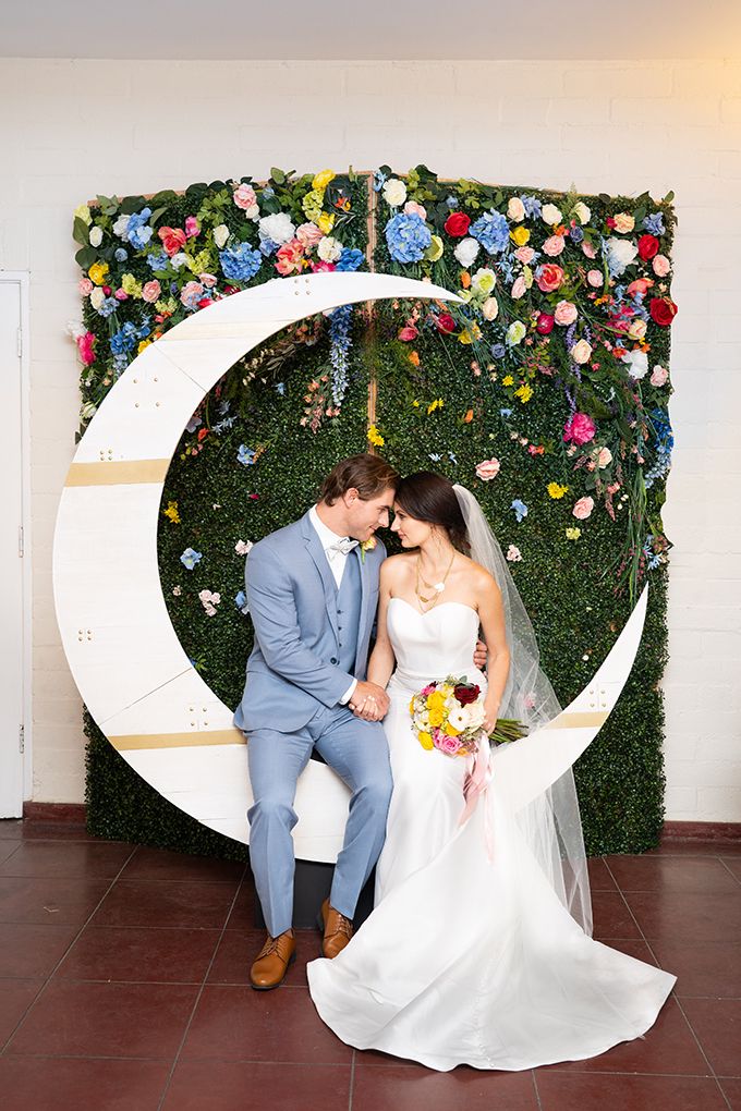 a bride and groom sitting on the moon in front of a wall with artificial flowers