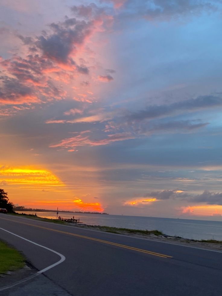 the sun is setting over the ocean with clouds in the sky and some grass on the side of the road