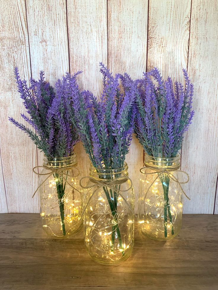 three mason jars filled with lavender flowers on top of a wooden table next to string lights