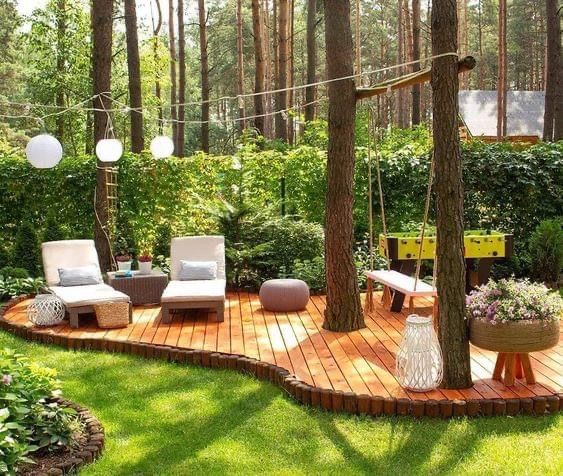 a wooden deck in the middle of a lush green yard with hanging lights and chairs