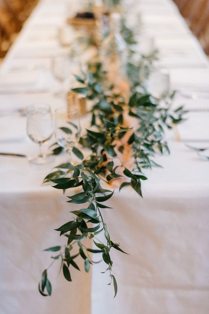 the table is set with white linens and greenery on it, along with wine glasses