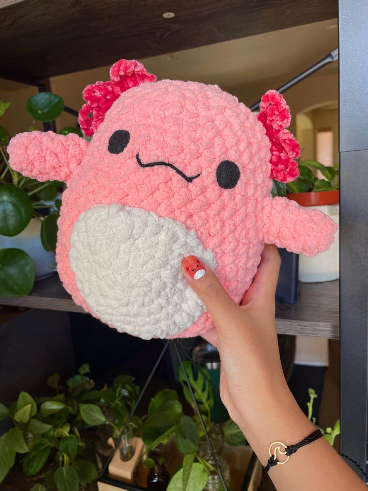 a hand holding a pink and white stuffed animal in front of a potted plant