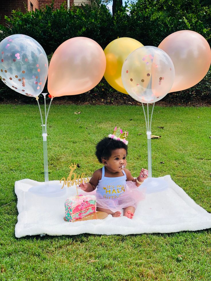 a baby sitting on a blanket in the grass with balloons behind her and a birthday cake