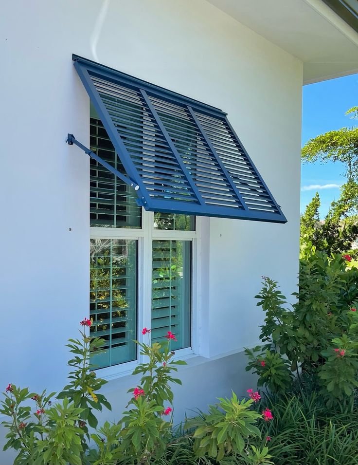 an open window with blue shutters on the side of a white building next to plants and flowers