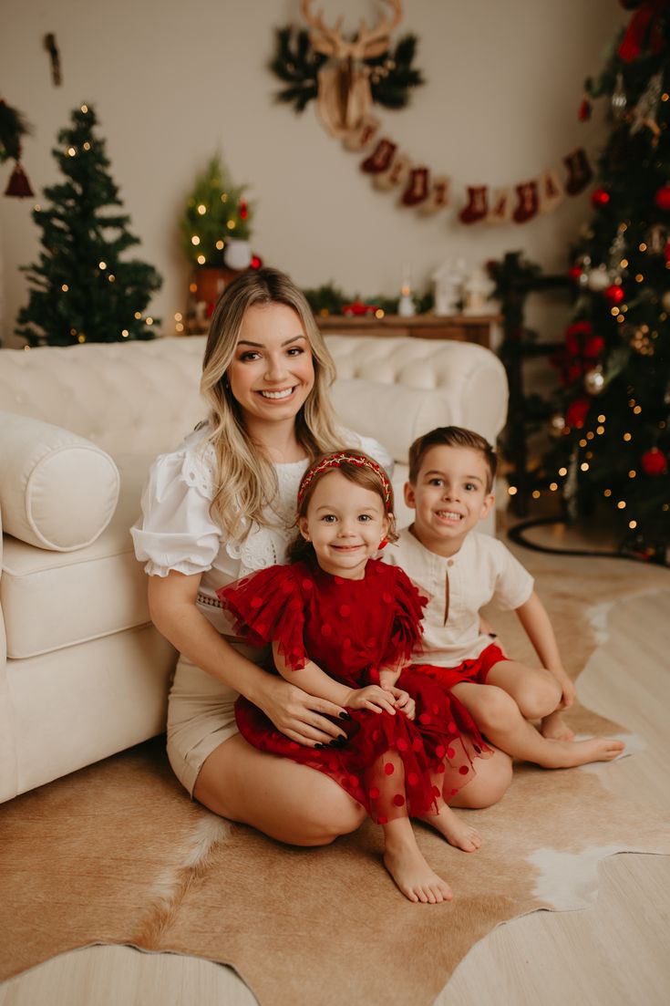 a mother and her two children sitting on the floor in front of christmas tree decorations