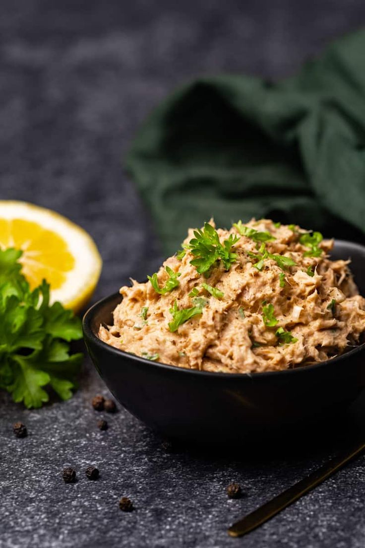 a black bowl filled with tuna salad next to a lemon slice and some parsley