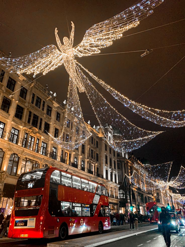 a red double decker bus driving down a street next to tall buildings covered in christmas lights